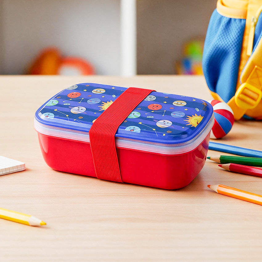 A red lunch box with a blue lid printed with cartoon planets and stars, secured with a red elastic strap, sitting on a light wood desk with school supplies nearby.
