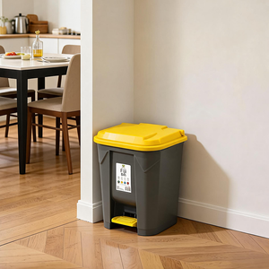 A gray and yellow plastic waste bin with a foot pedal, placed in the corner of a kitchen or dining room on a wood floor against a beige wall.